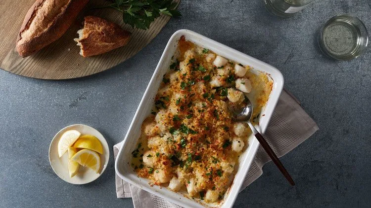 Martha Stewart baked scallops in a white baking dish topped with golden panko and parsley, next to lemon wedges and crusty bread