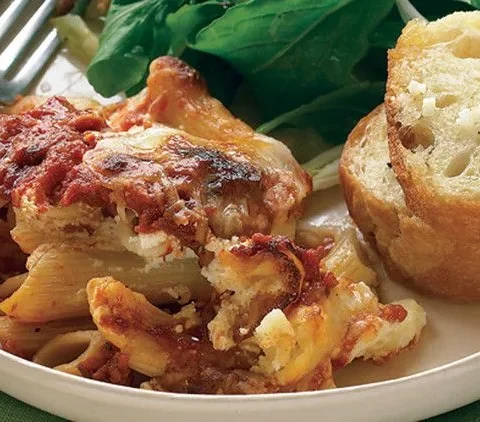 Martha Stewart baked ziti with browned cheese and tomato sauce served with garlic bread and a green salad on a white plate