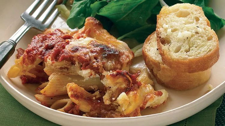 Martha Stewart baked ziti with browned cheese and tomato sauce served with garlic bread and a green salad on a white plate