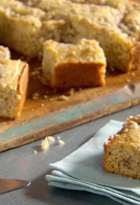 Martha Stewart banana cake squares with brown sugar crumb topping on a wooden board and a single piece on a pale blue napkin