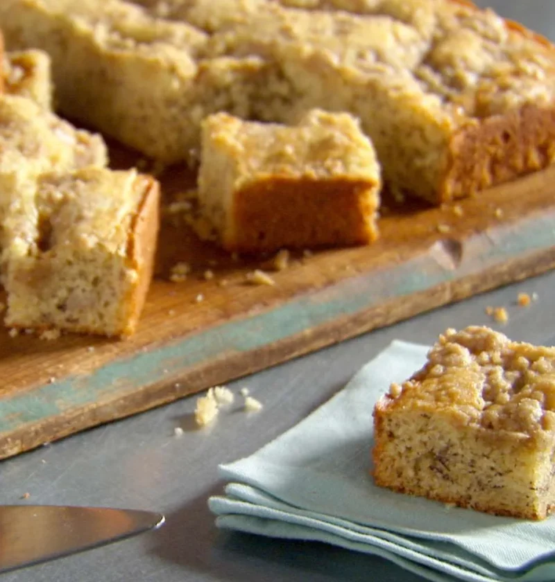 Martha Stewart banana cake squares with brown sugar crumb topping on a wooden board and a single piece on a pale blue napkin