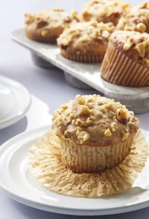 Martha Stewart banana nut muffins topped with chopped walnuts served on white plates with cups of coffee
