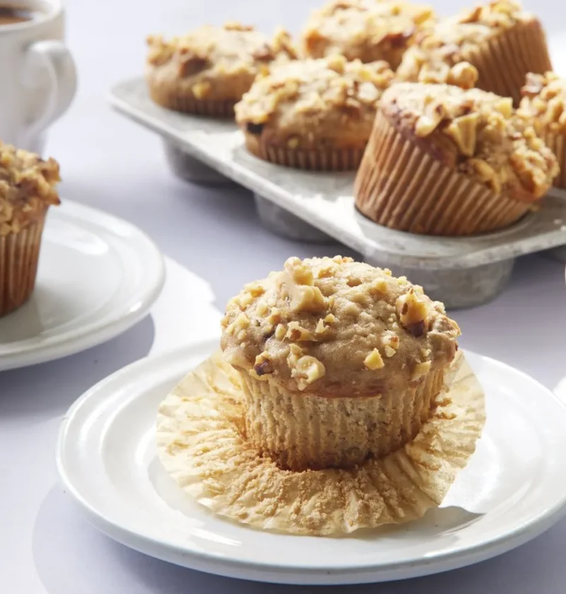 Martha Stewart banana nut muffins topped with chopped walnuts served on white plates with cups of coffee