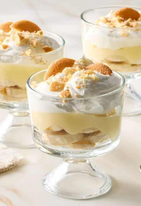 Martha Stewart banana pudding cups in glass pedestal dishes topped with whipped cream and crumbled vanilla wafer cookies