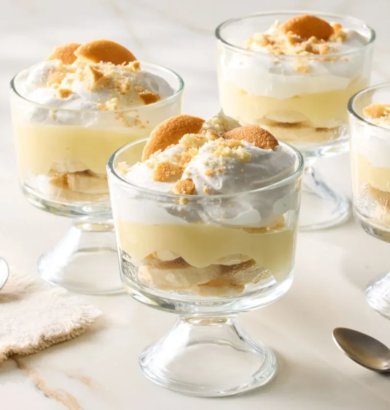 Martha Stewart banana pudding cups in glass pedestal dishes topped with whipped cream and crumbled vanilla wafer cookies