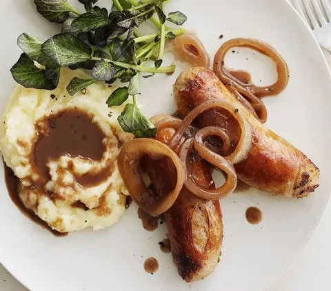 Martha Stewart bangers and mash with browned sausages, onion gravy and cheesy mashed potatoes beside watercress on a white plate