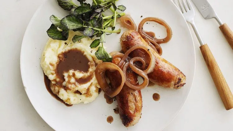 Martha Stewart bangers and mash with browned sausages, onion gravy and cheesy mashed potatoes beside watercress on a white plate
