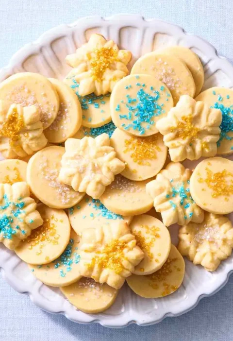 Martha Stewart basic butter cookie dough shaped into round and spritz cookies with blue and gold sprinkles on a white scalloped plate