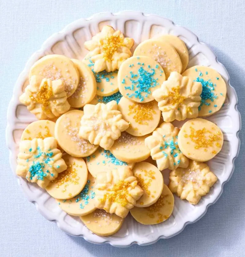 Martha Stewart basic butter cookie dough shaped into round and spritz cookies with blue and gold sprinkles on a white scalloped plate