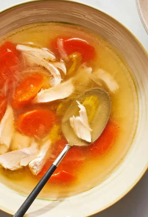 Martha Stewart basic chicken soup in a shallow bowl with shredded chicken, sliced carrots and a spoon in golden broth