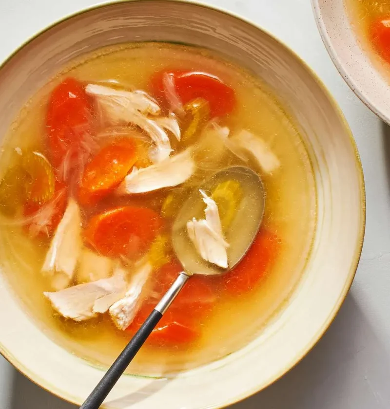 Martha Stewart basic chicken soup in a shallow bowl with shredded chicken, sliced carrots and a spoon in golden broth