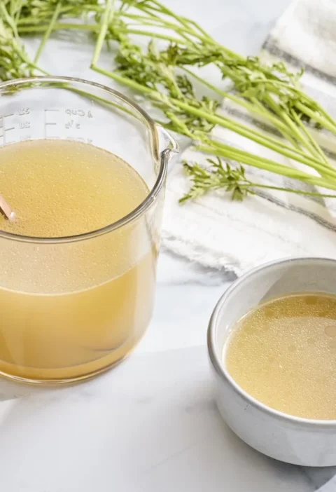 Martha Stewart basic chicken stock, clear golden broth in a glass measuring cup beside a small bowl and fresh carrots