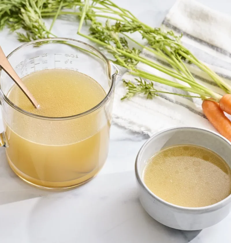 Martha Stewart basic chicken stock, clear golden broth in a glass measuring cup beside a small bowl and fresh carrots