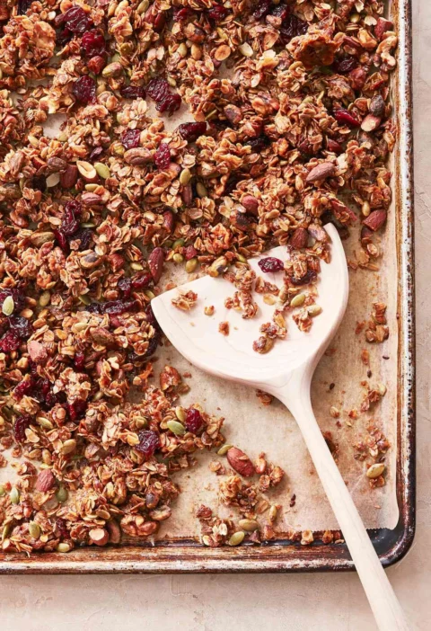 Martha Stewart basic granola with oats, nuts, pumpkin seeds and dried cranberries on a baking sheet with a wooden spatula
