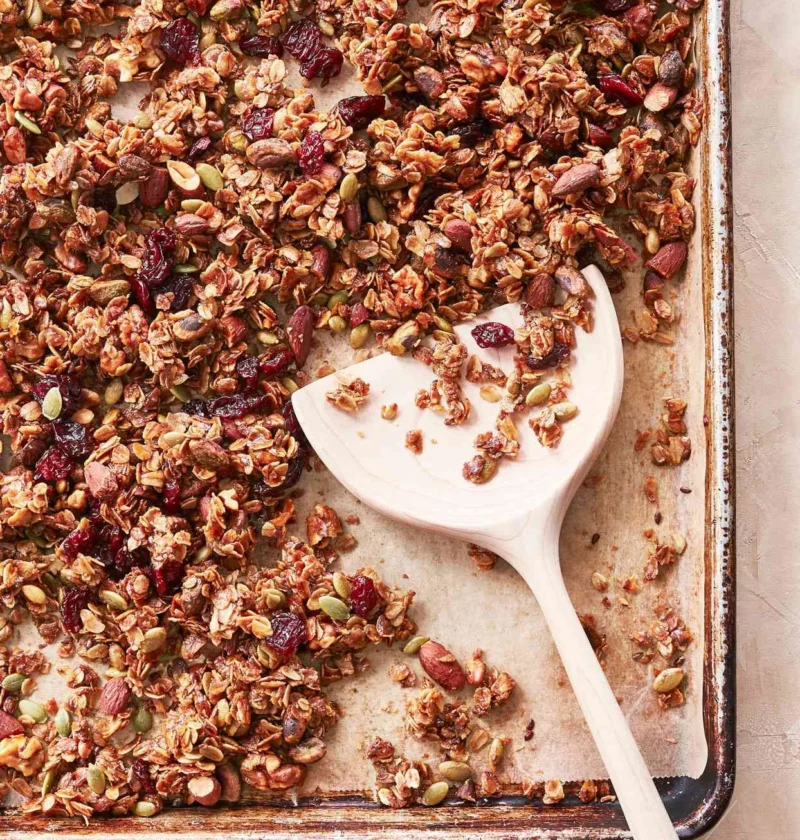 Martha Stewart basic granola with oats, nuts, pumpkin seeds and dried cranberries on a baking sheet with a wooden spatula