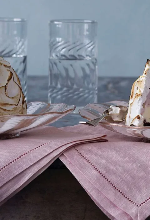 Two Baked Alaska desserts topped with toasted Italian meringue, one cut open to show pink ice cream layers, on glass plates with pink napkins