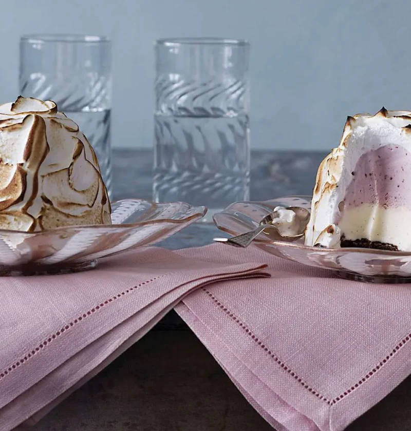 Two Baked Alaska desserts topped with toasted Italian meringue, one cut open to show pink ice cream layers, on glass plates with pink napkins