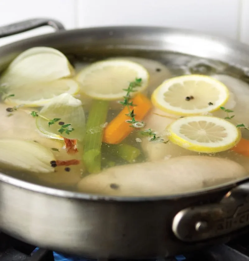 Martha Stewart basic poached chicken breasts simmering in a skillet with lemon slices, carrot, celery, onion and thyme