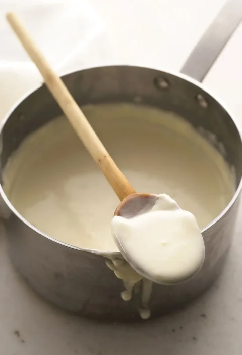 Martha Stewart bechamel sauce in a saucepan with a wooden spoon lifting up a thick, creamy spoonful