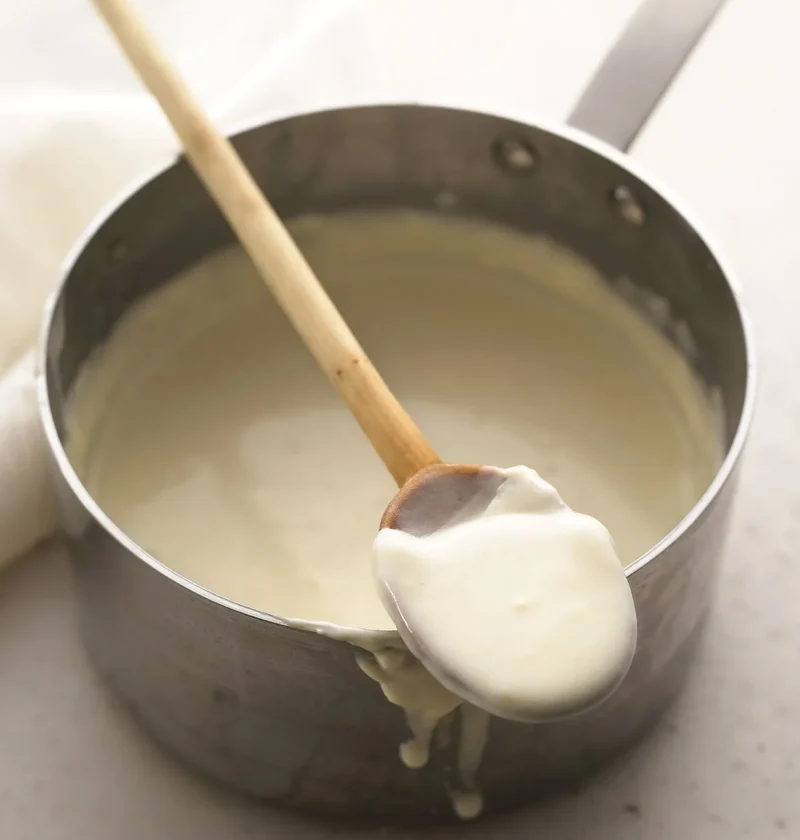 Martha Stewart bechamel sauce in a saucepan with a wooden spoon lifting up a thick, creamy spoonful