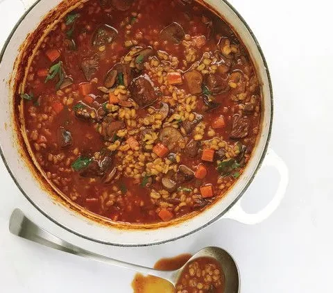 Martha Stewart beef and barley soup with beef cubes, barley, carrots and mushrooms in a white Dutch oven with a spoon beside it