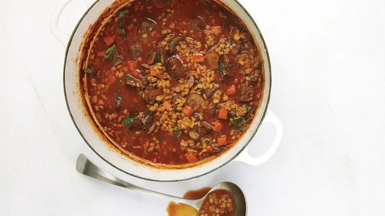 Martha Stewart beef and barley soup with beef cubes, barley, carrots and mushrooms in a white Dutch oven with a spoon beside it