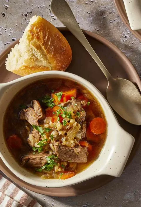 Martha Stewart beef barley soup with beef chunks, carrots, barley and parsley in a cream bowl with crusty bread
