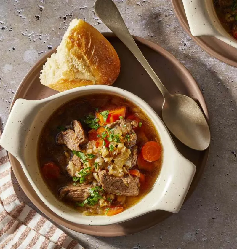 Martha Stewart beef barley soup with beef chunks, carrots, barley and parsley in a cream bowl with crusty bread