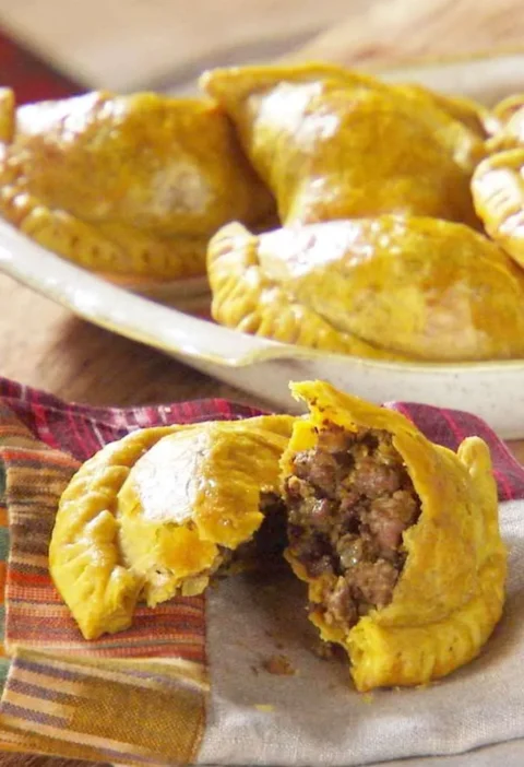 Martha Stewart beef patties, golden Jamaican half-moon pastries on a plate, one broken open to show the spiced beef filling next to a bottle of hot sauce