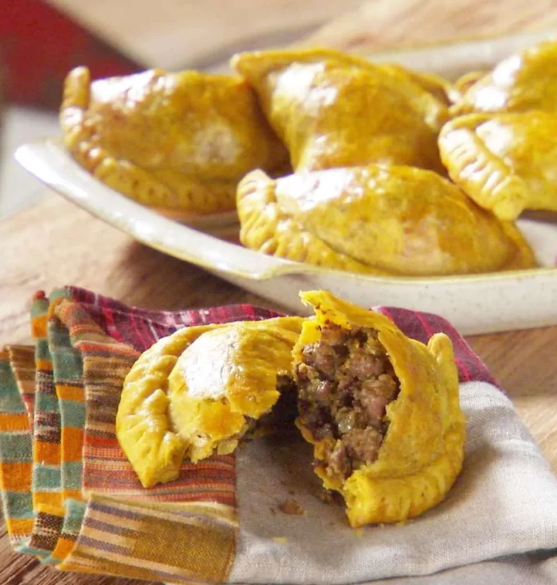 Martha Stewart beef patties, golden Jamaican half-moon pastries on a plate, one broken open to show the spiced beef filling next to a bottle of hot sauce