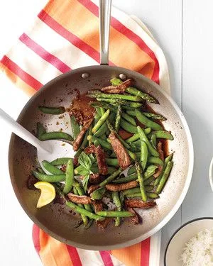 Martha Stewart beef, snap pea, and asparagus stir-fry in a skillet with basil and a lemon wedge next to a bowl of rice