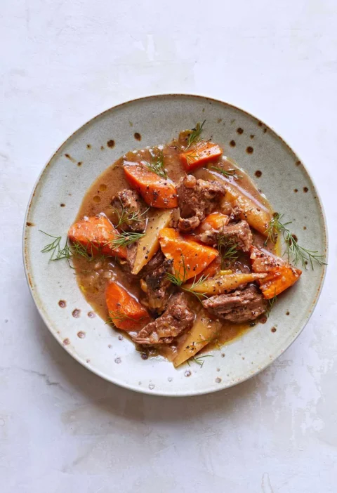 Martha Stewart beef stew with sweet potatoes, carrots and parsnips in a light brown broth, topped with fresh dill in a speckled bowl