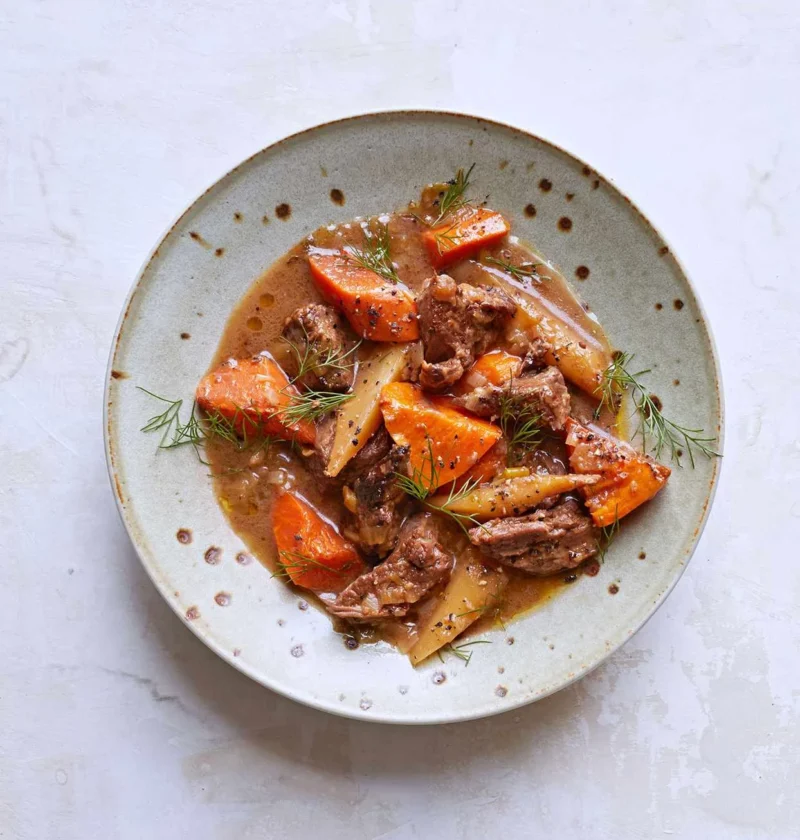 Martha Stewart beef stew with sweet potatoes, carrots and parsnips in a light brown broth, topped with fresh dill in a speckled bowl