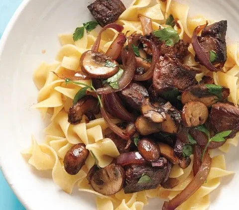Martha Stewart beef stroganoff stir-fry with seared steak, red onion slices, cremini mushrooms and parsley over egg noodles in a white bowl