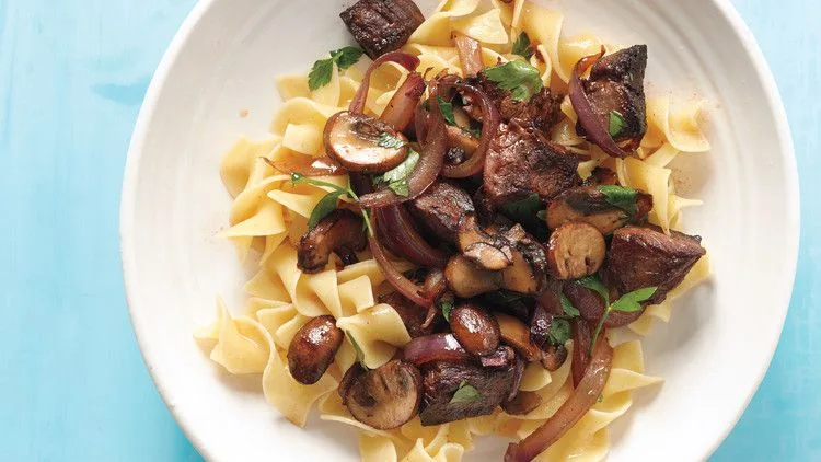 Martha Stewart beef stroganoff stir-fry with seared steak, red onion slices, cremini mushrooms and parsley over egg noodles in a white bowl