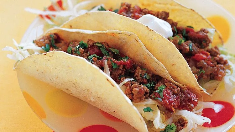 Martha Stewart beef tacos in corn tortillas filled with saucy ground beef, shredded lettuce, cilantro and sour cream on a colorful plate