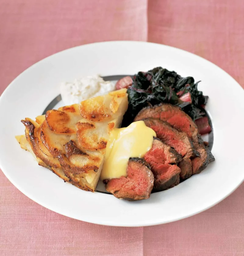 Sliced medium-rare Martha Stewart beef tenderloin served with bearnaise sauce, potato gratin and sauteed greens on a white plate