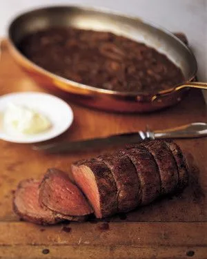 Sliced Martha Stewart beef tenderloin with shallot mustard sauce on a wooden cutting board next to a skillet of sauce