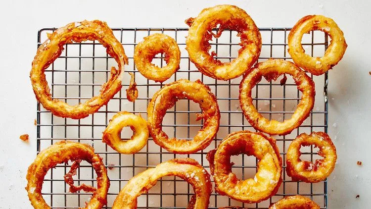 Martha Stewart beer-battered onion rings fried golden brown and cooling on a wire rack with flaky sea salt