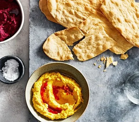 Martha Stewart beet-and-tahini dip in a small white bowl next to a yellow dish of hummus with harissa oil, flatbread shards and a salt dish