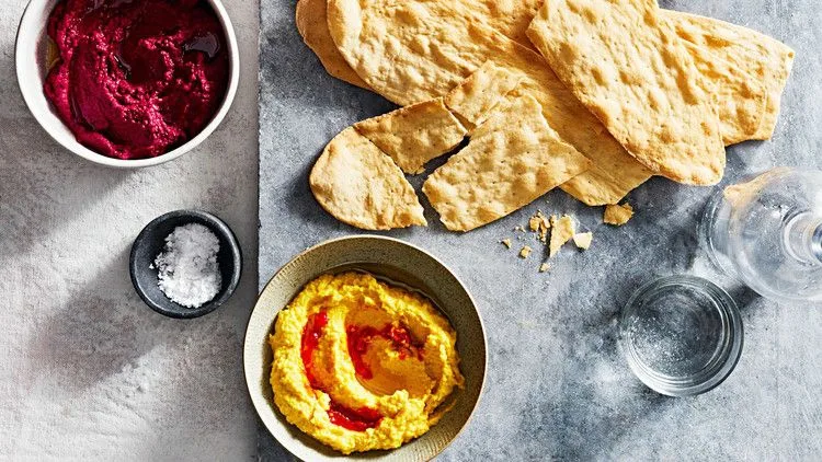 Martha Stewart beet-and-tahini dip in a small white bowl next to a yellow dish of hummus with harissa oil, flatbread shards and a salt dish