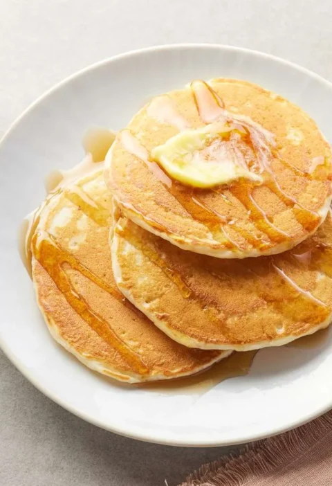 Martha Stewart best buttermilk pancakes stacked on a white plate with melting butter and syrup beside a glass of orange juice