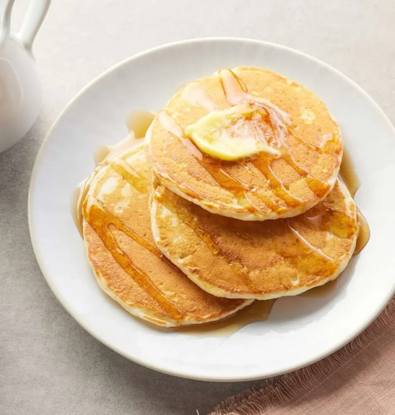 Martha Stewart best buttermilk pancakes stacked on a white plate with melting butter and syrup beside a glass of orange juice