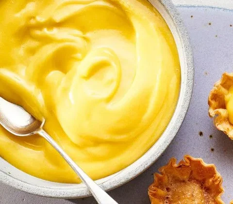 Martha Stewart best lemon curd in a gray bowl with a spoon next to small tartlet shells filled with lemon curd