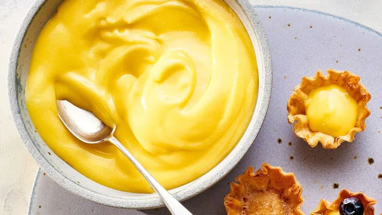 Martha Stewart best lemon curd in a gray bowl with a spoon next to small tartlet shells filled with lemon curd