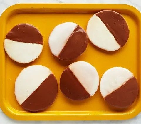 Six Martha Stewart black and white cookies iced half vanilla and half chocolate on a yellow tray