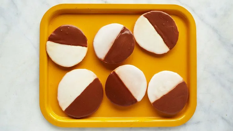 Six Martha Stewart black and white cookies iced half vanilla and half chocolate on a yellow tray