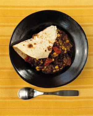 Martha Stewart black-bean-and-corn stew in a black bowl with a folded flour tortilla and a spoon on a yellow striped cloth