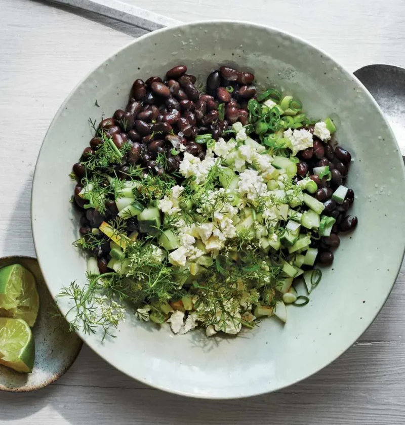 Martha Stewart black bean, cucumber, and feta salad topped with crumbled feta, diced cucumber and fresh herbs in a pale green bowl with lime halves