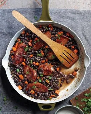 Martha Stewart black beans and sausage in a white skillet with browned kielbasa, diced carrots and parsley beside a wooden spatula